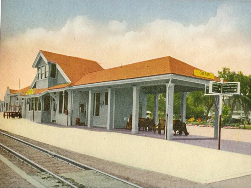 An older image of a train depot with patrons waiting on park benches